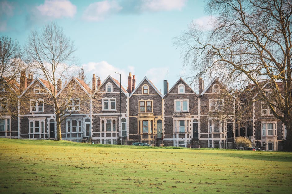 The image depicts a row of traditional Victorian-style terraced houses made of grey stone with white window frames and decorative elements, set against a partly cloudy sky in Finsbury Park. In the foreground, there is a well-maintained grassy area with a few leafless trees, indicating a season of late autumn or winter. The houses are positioned behind a low fence, and in front of the houses, there is a paved sidewalk or street with a few cars parked along the curb. The scene is illuminated by natural daylight, with the shadows of the trees cast on the grass. While the image does not show any moving activities directly related to house removals, the setting indicates an urban residential area where professional removal services, like those provided by Man With a Van Finsbury Park, may be involved in a home relocation or furniture transport process. The overall scene is calm and orderly, capturing the environment where packing, loading, and transportation activities for a move could take place.