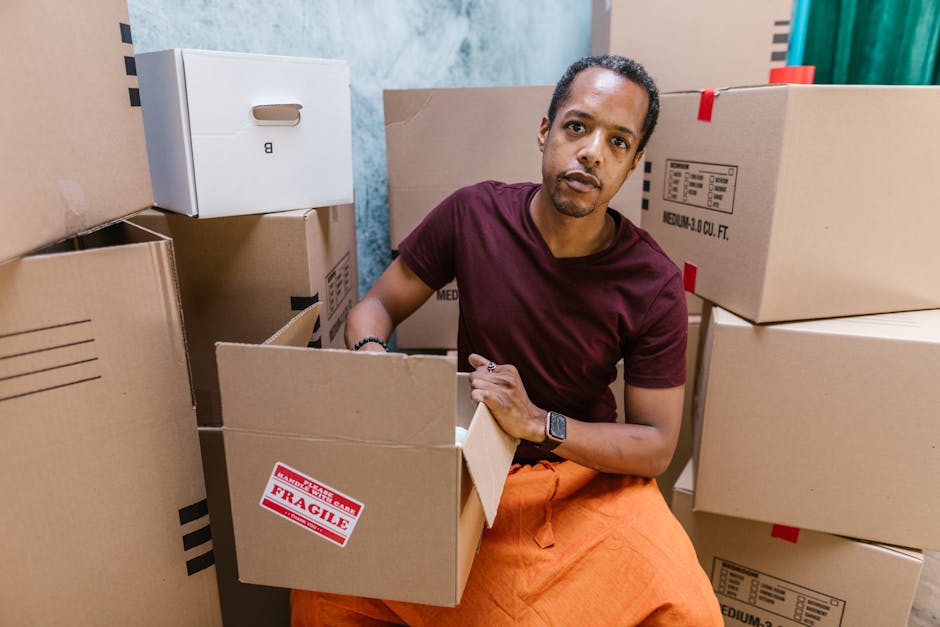 A man wearing a maroon t-shirt and orange pants sits inside a room filled with stacked cardboard boxes, some of which are labeled as fragile. He is holding open a cardboard box with one hand, while the other hand rests on the edge of the box, with several other boxes surrounding him inside the property. The boxes vary in size and are sealed with packing tape, indicating they are packed for moving. The environment appears to be a home or storage area, with a blue wall and some indoor decorative elements visible in the background. The setting reflects the packing and loading process involved in house removals or furniture transport services provided by Man With a Van Finsbury Park, highlighting aspects of household packing and the careful handling of items during a relocation.