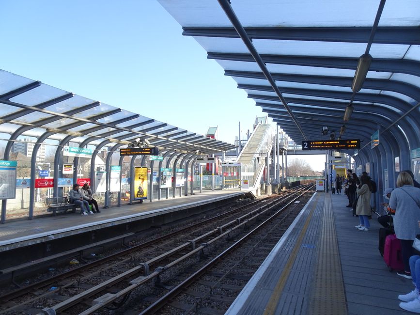 A view of Finsbury Park station platform featuring a covered waiting area with glass and metal roofing, illuminated digital service information boards, and seating with a few passengers sitting. In the background, there are multiple railway tracks extending into the distance, with a station staircase leading to an overpass. On the right side, several people are standing and sitting while waiting for their train, some with luggage and backpacks. The platform surface is paved with tactile paving strips near the edge for accessibility. The clear sky and daylight illuminate the station environment, which includes advertisements and safety notices posted along the shelter, reflecting typical elements of a busy urban train station, suitable for home relocation or furniture transport logistics where efficient passenger and cargo movements are involved.
