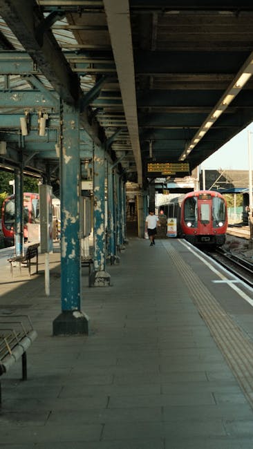 A photograph of an underground London underground station platform near Finsbury Park, showing a partially visible red train approaching from the distance. The platform is tiled with beige and brown tiles, and features safety markings including a yellow line along the edge and textured tactile paving for visually impaired passengers. Along the wall, there are several posters and advertisements, including a prominent blue poster for Moneybox. Accessible signage, including a blue wheelchair symbol and an arrow pointing towards the stairs, is visible on the tiled wall near the platform entrance, which has a staircase with black metal steps and a handrail. The ceiling consists of metal panels with integrated lighting fixtures, providing illumination for the platform area. This setting is relevant for home relocation activities in central London, where furniture transport and packing are coordinated with access and safety considerations for moving day.