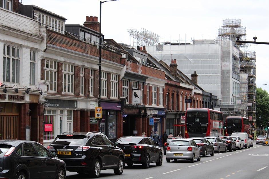 The image depicts a busy urban street scene in Finsbury Park, with a row of brick-fronted terraced houses lining the pavement. Several vehicles, including a black car, a silver car, and a Lexus SUV, are parked along the roadside. Behind the parked cars, a group of pedestrians is walking on the sidewalk. In the background, there are two red double-decker buses, with route numbers 343 and 142, stopped at a bus stop. The street scene also features a construction site on the right side, covered in scaffolding and white protective sheeting, indicating ongoing building work. The overcast sky creates diffused lighting over the scene. Occasionally visible are shopfronts and signage, including a store with a purple awning, contributing to the lively commercial atmosphere typical of a neighbourhood involved in home relocation and moving services. The overall setting combines elements of daily city life with ongoing infrastructure projects, reflecting an environment where furniture transport, packing, and moving logistics are common.