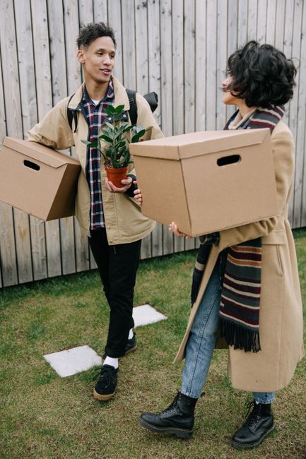 Two individuals engaged in a home relocation process outdoors, standing on a grassy area adjacent to a wooden fence. The person on the left, a young man with short curly hair, is dressed in black trousers, a checkered shirt layered with a beige jacket, and is carrying a cardboard box in each hand. The person on the right, a woman with dark, curly hair, is wearing a long beige coat, a striped scarf, and black boots, and is holding a medium-sized cardboard box. Between them, a small potted plant with green leaves is being handed over, indicating careful handling of belongings. The scene suggests the final stage of packing and moving, with visible packing materials and prepared boxes, characteristic of professional removals services by [COMPANY_NAME], emphasizing organized furniture transport and loading procedures in an outdoor environment, ready for a house move.