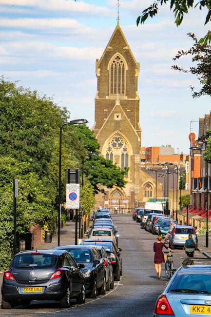 Image showing a street scene near Seven Sisters Road in Finsbury Park, with a prominent historic church featuring a tall, pointed spire and large stained glass windows in the background. The street is lined with parked cars on both sides, including sedans and hatchbacks, with some vehicles partially visible as they extend out of view. Green trees and foliage are visible along the sidewalk, providing shade and contrast to the urban environment. In the foreground, a person wearing a maroon coat and black shoes is walking beside a bicycle, with another individual in casual clothing nearby. Street lamps and signage are also present along the pavement. This scene depicts a typical urban setting that could be part of a home relocation or furniture transport process, with potential for loading vehicles or moving boxes in a residential or commercial area, consistent with the services of Man With a Van Finsbury Park.