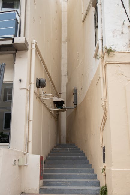 A close-up view of a staircase in an interior setting, with dark, worn steps and a wooden handrail attached to a white tiled wall. The staircase appears narrow, and part of a green protective covering